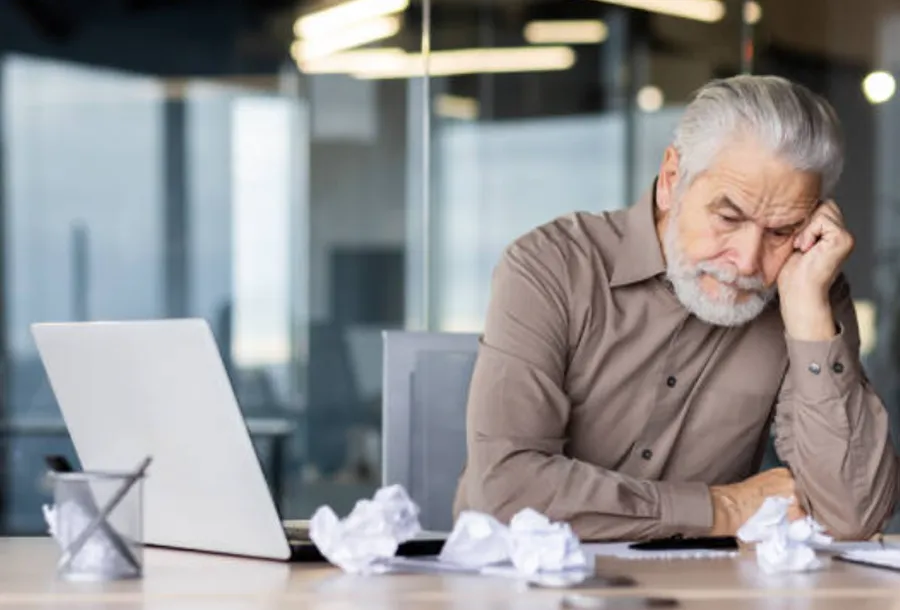 A frustrated businessman sitting at laptop with crumbled papers scattered on desk